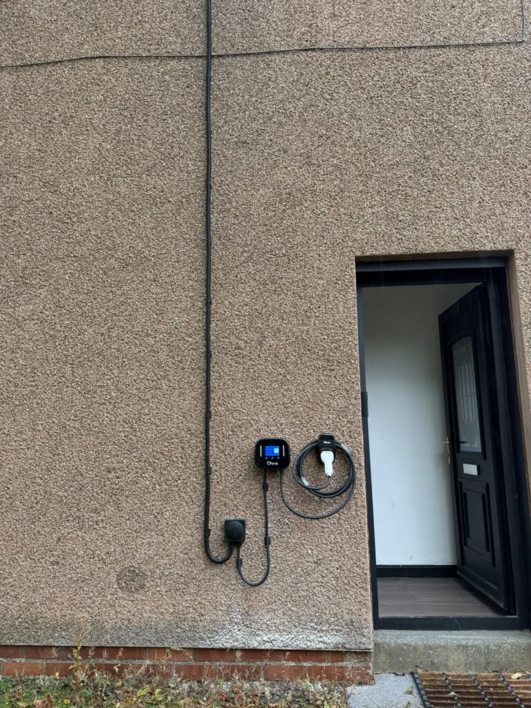 Ohme Home Pro EV charger installed beside a doorway in West Calder, with its screen, coiled charging cable and external cable route visible