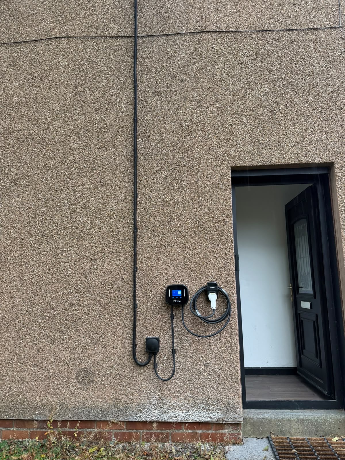 Ohme Home Pro EV charger installed beside a doorway in West Calder, with its screen, coiled charging cable and external cable route visible